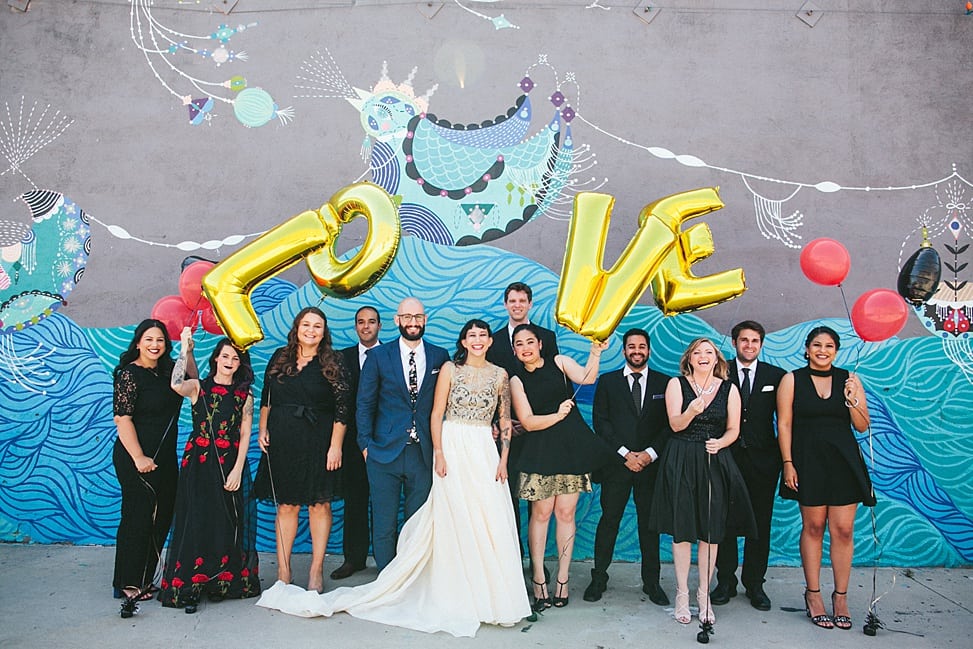 A group stand beside a wedding couple for a formal photo in front of a colorful graffitied wall. The wedding party holds gold letter balloons that spell LOVE and red latex balloons.