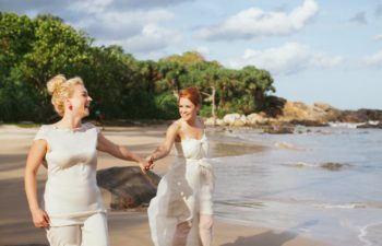 Two women in wedding dresses hold hands on a beach, smiling