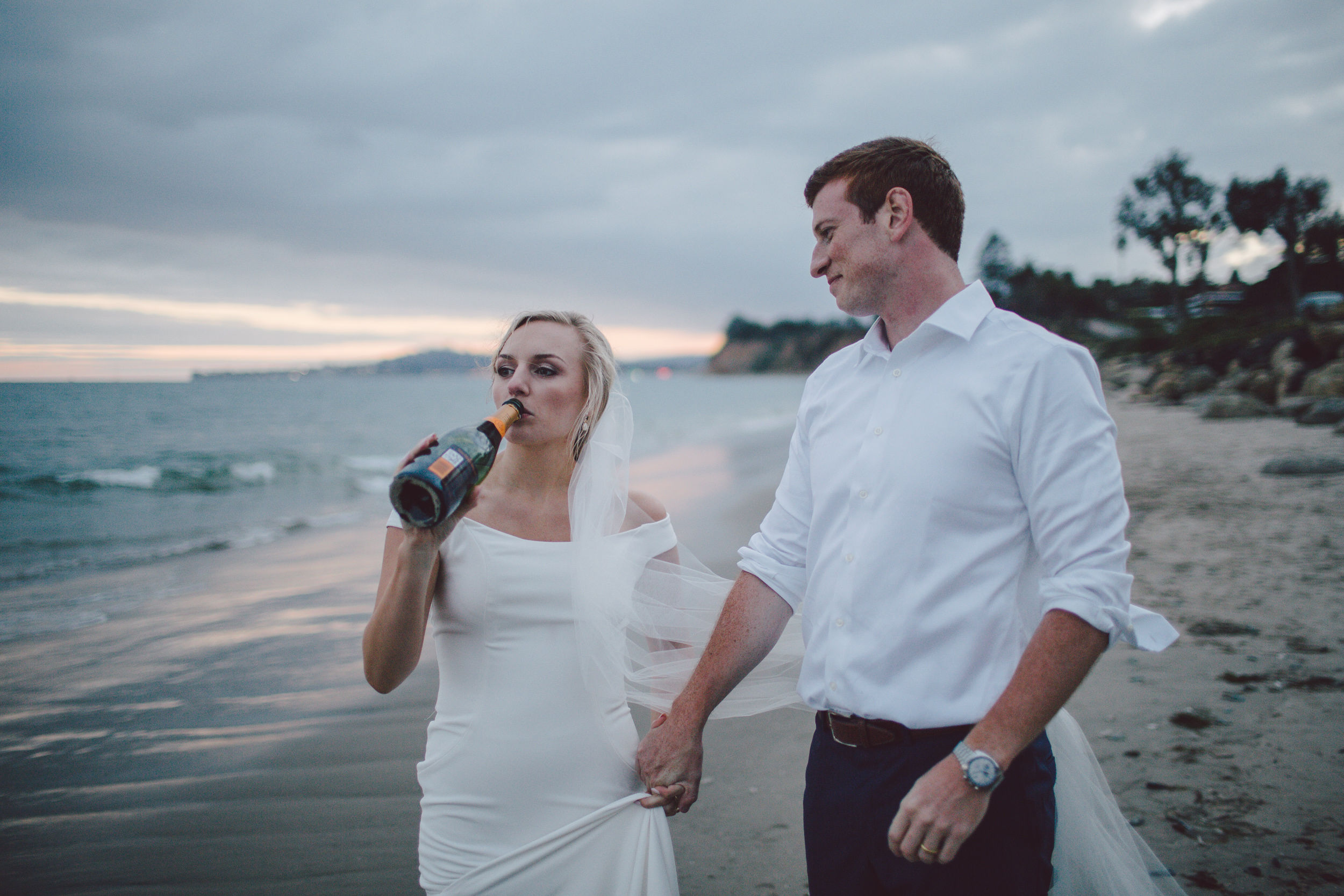 A couple walk along a beach as one of them drinks from a champagne bottle