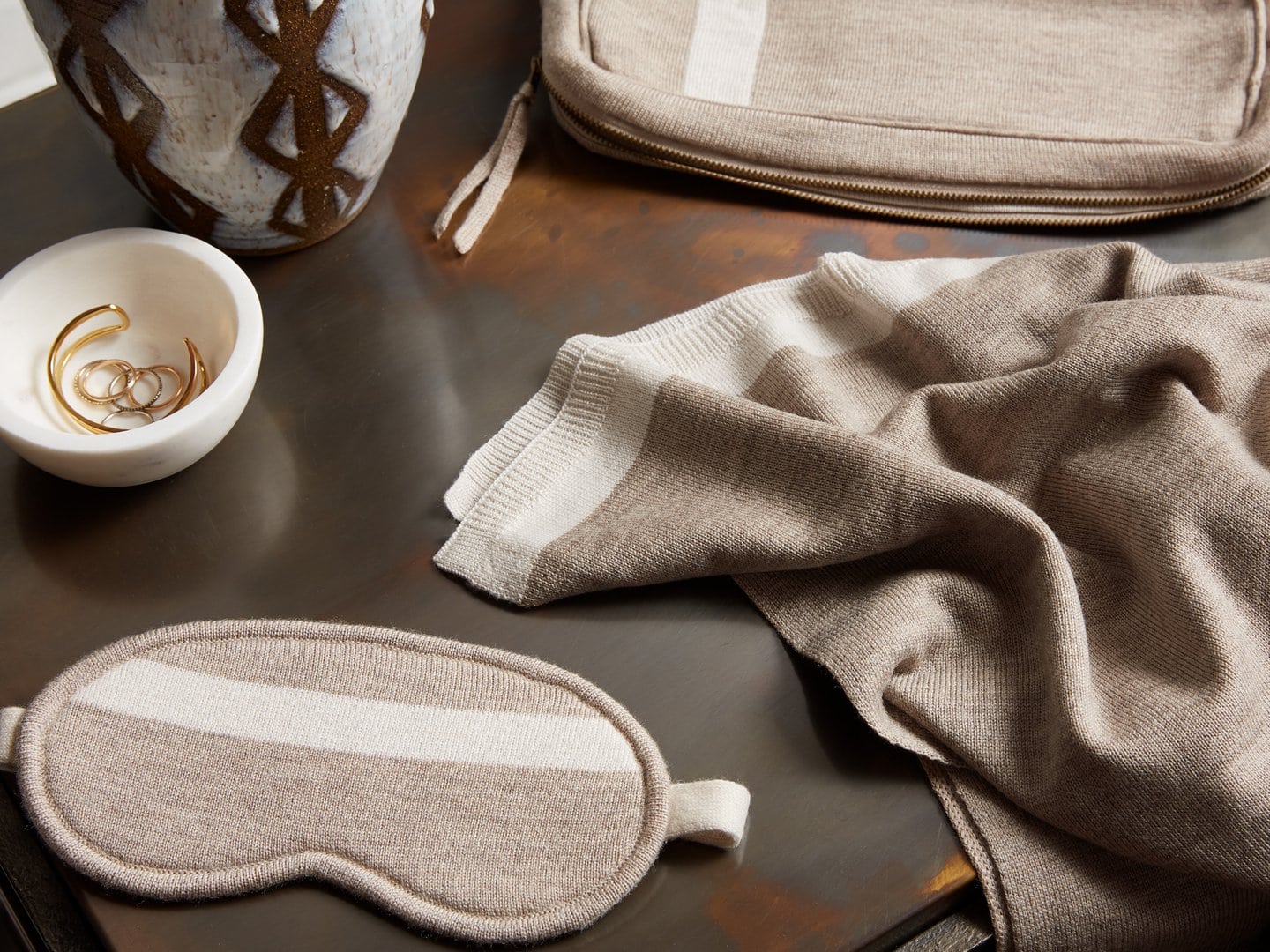 grey and white knit travel blanket and eye mask set on a table next to a white bowl holding gold jewelry