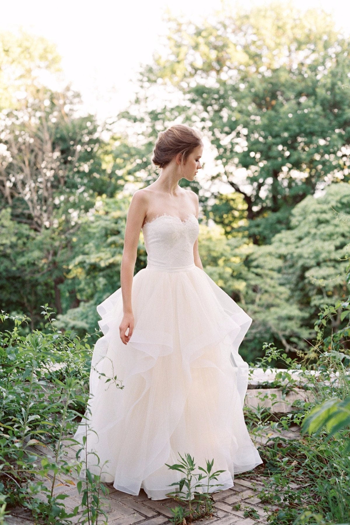 a woman wears a strapless pale blush dress with a sweetheart neckline, corset style bodice, and tiered tulle skirt