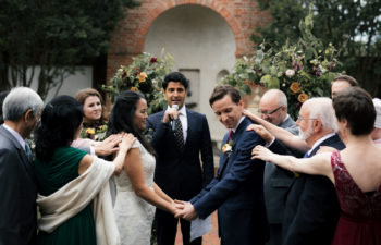 Couple holding hands at wedding ceremony in front of officiant, surrounded by family placing hands on them