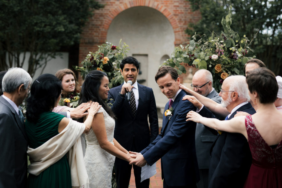 Couple holding hands at wedding ceremony in front of officiant, surrounded by family placing hands on them