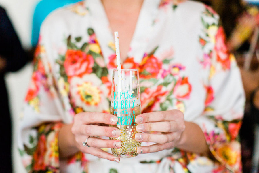 woman in flower robe holding a gold-spotted stemless champagne glass that reads "pop the bubbly I'm getting a hubby" in teal script, with a paper straw