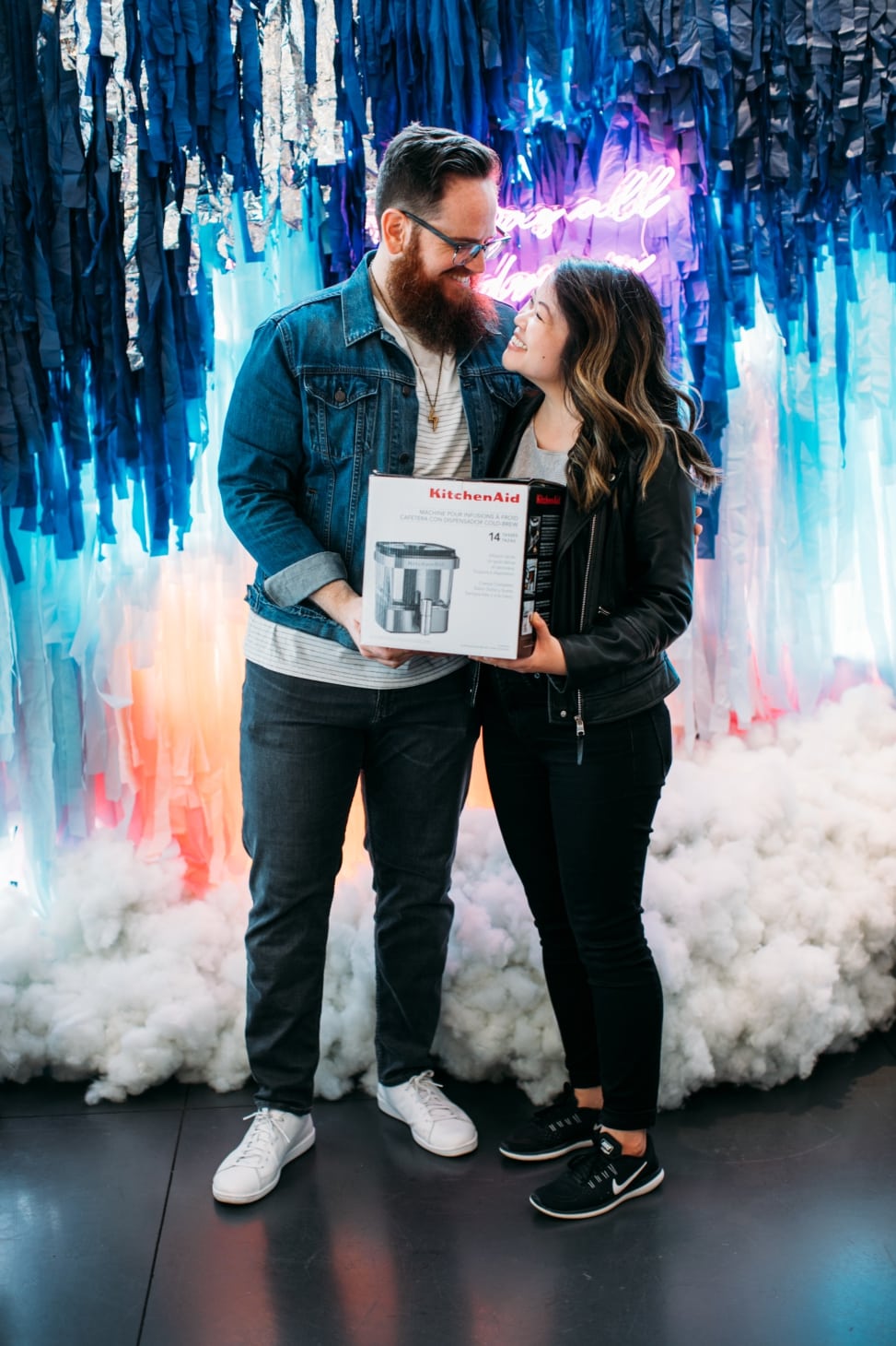 A couple stands together in front of a shimmery backdrop with clouds holding a boxed KitchenAid appliance at the crate and barrel san francisco private registry event with a practical wedding