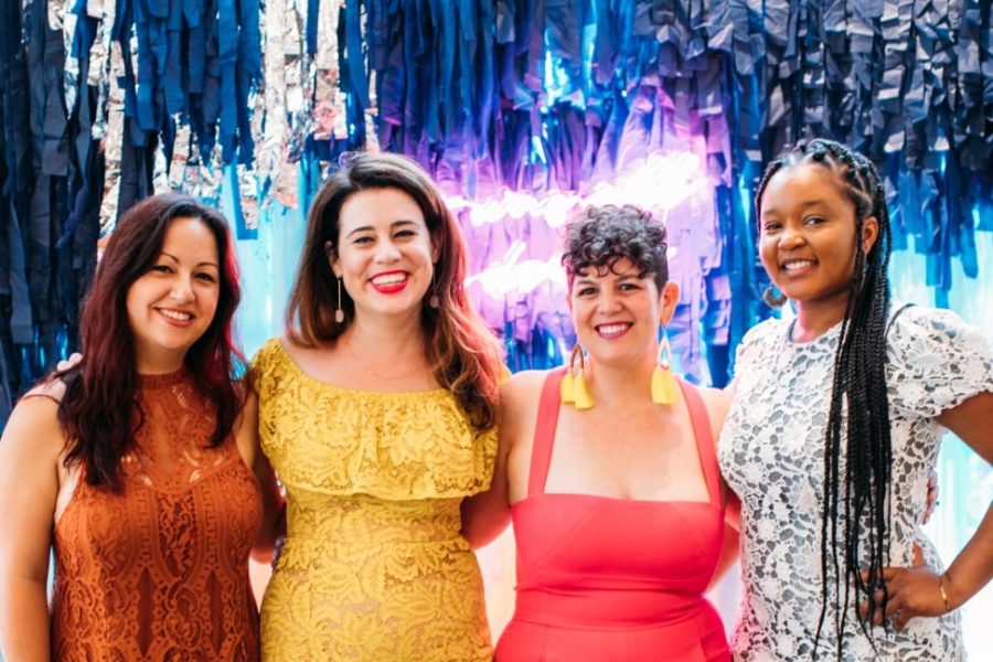 Four women in party clothes stand in front of a waterfall fringe and neon light installation at a party