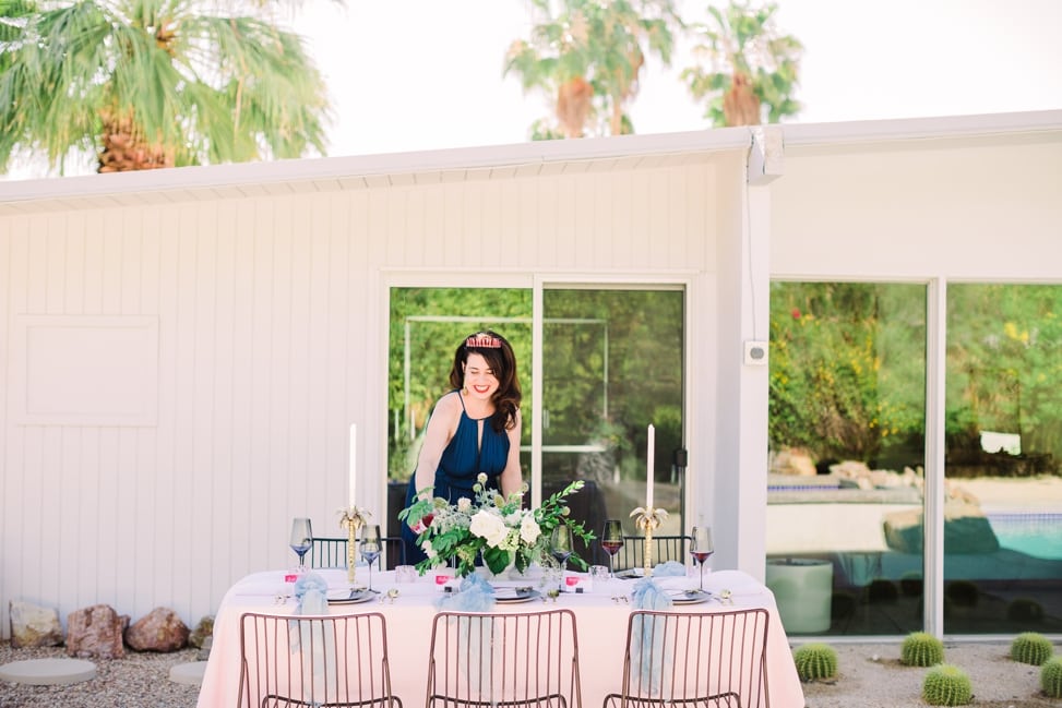 a woman in a sundress and tiara prepares a dinner table outdoors