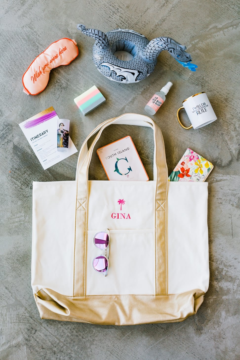 a personalized embroidered tote bag that has a palmtree and reads "Gina" in pink, with sunglasses, a dragon cupholder pool floaty, a sleep mask that reads "Wish you were here," a gold trimmed mug, and other items