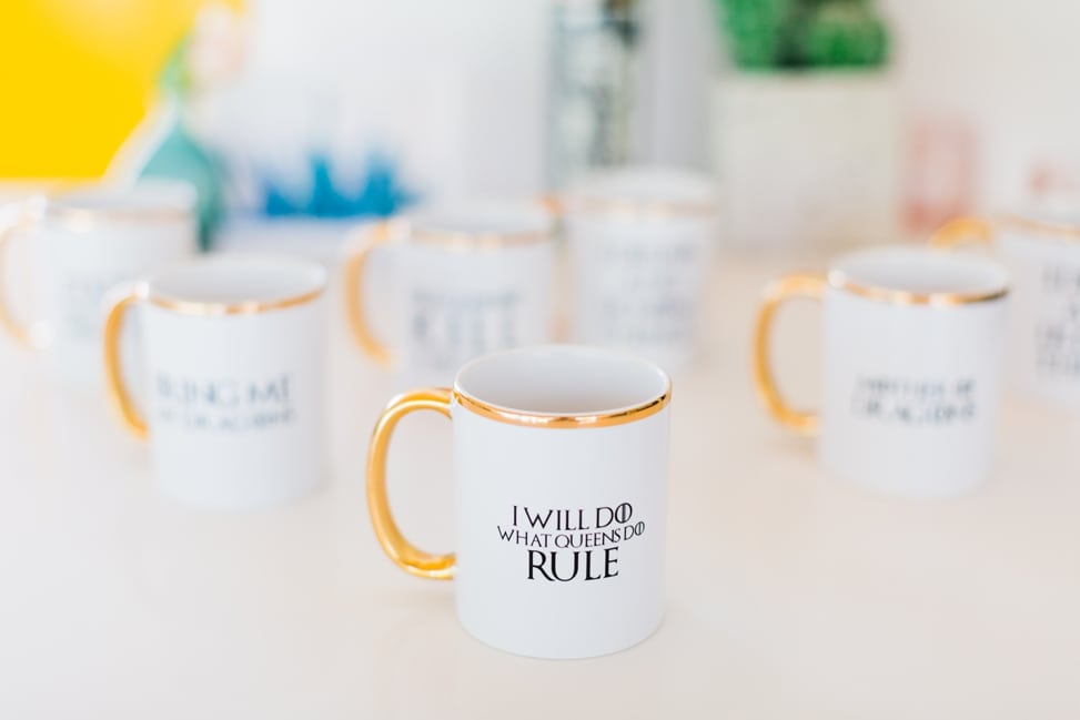 a table of white mugs with gold trim with different phrases printed on them; the closest reads "I will do what queens do / RULE"