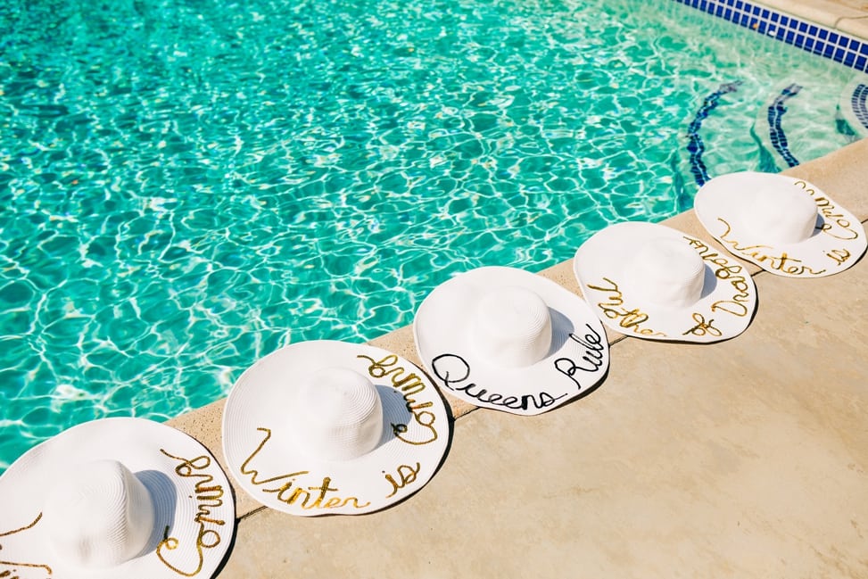 Five white sun hats line the edge of a pool and read Queens Rule (in black), Mother of Dragons (in gold), and 3 read Winter is Coming (also in gold)