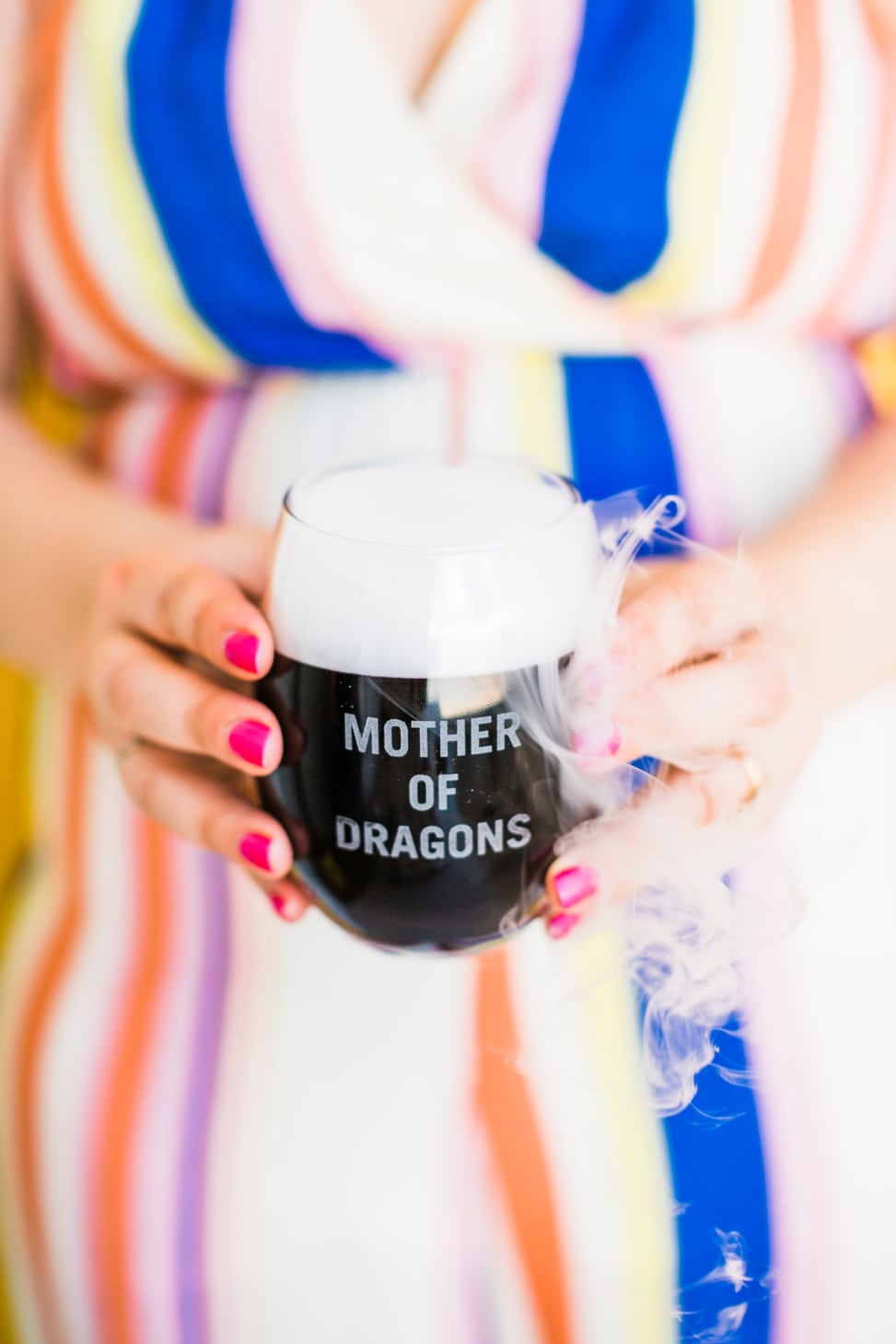 A woman in a brightly striped jumpsuit holds a stemless wineglass that reads "Mother of Dragons" and holds a black liquid that is smoking out of the top