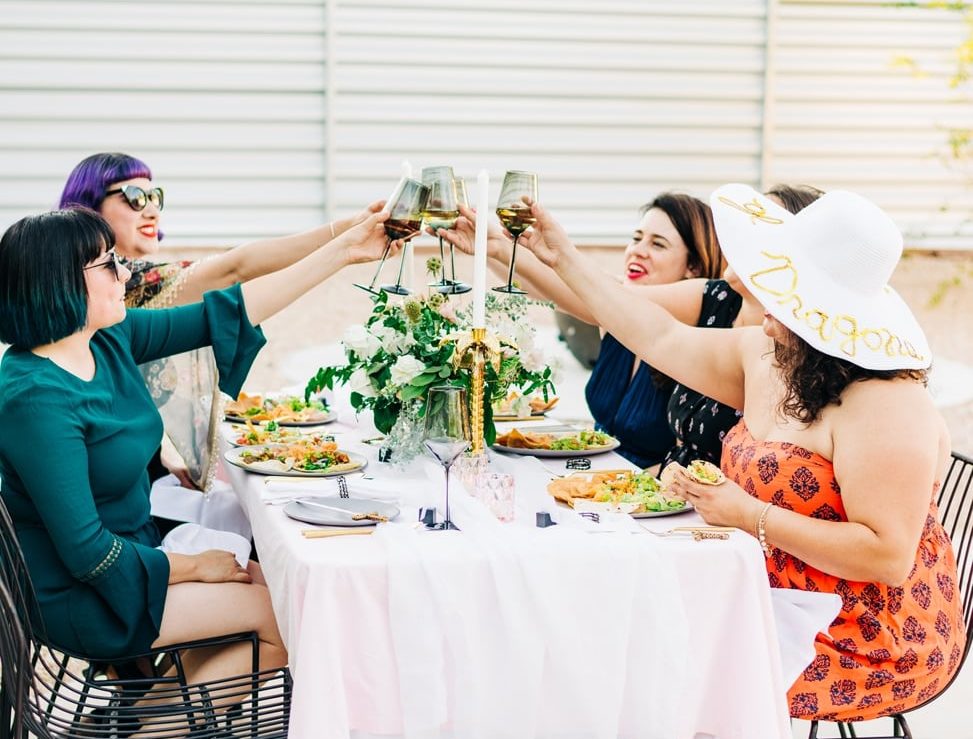 Women toasting at a table with a game of thrones inspired tablescape with pink tablecloth and blue silk napkins with palm tree candleholders and sword letter openers