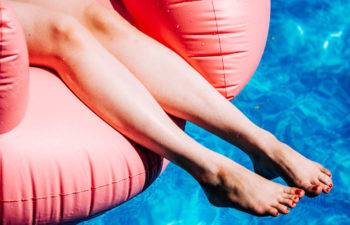 close up of women's legs and feet hanging off of pink flamingo floatie in blue pool