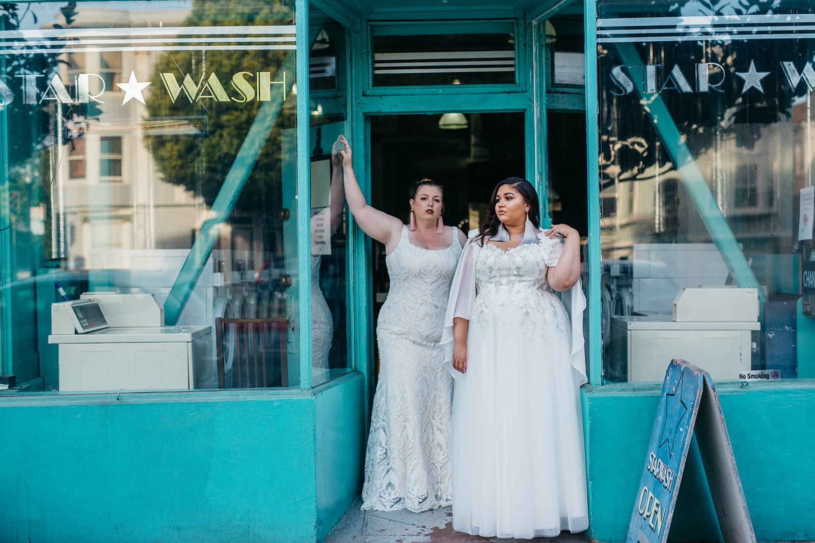 Two women in APW x Lace & Liberty plus size wedding dresses in the doorway of a teal-painted laundromat in an image by Studio XIII Photography