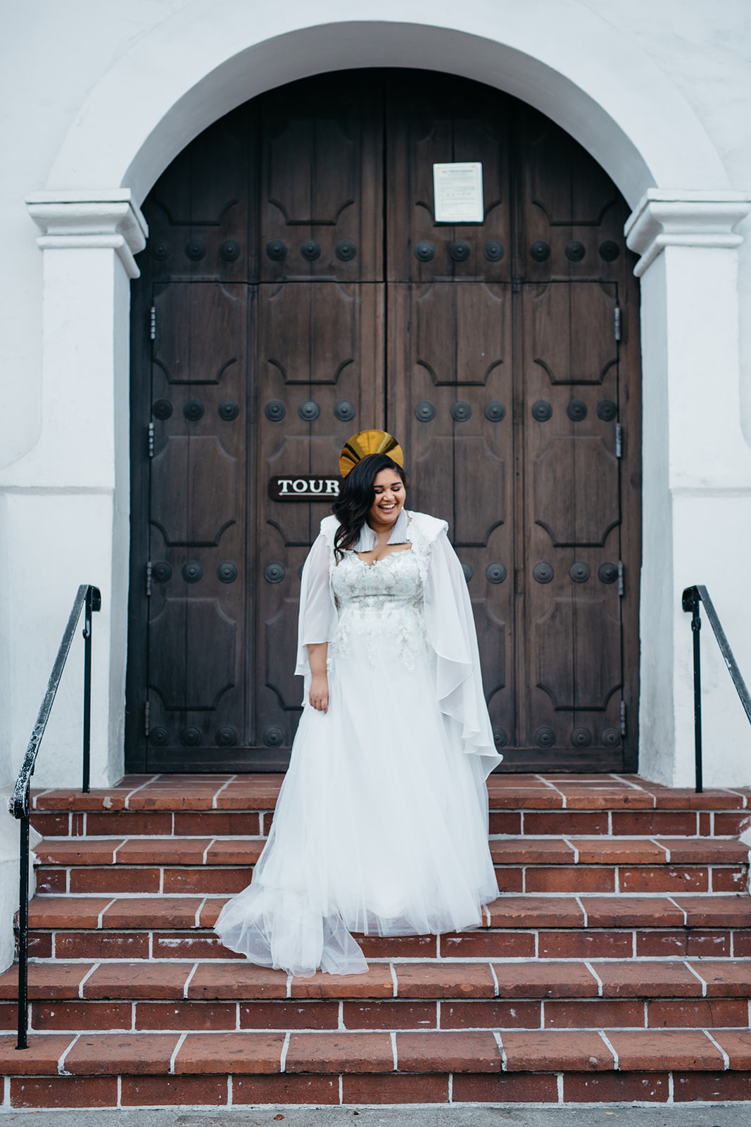 A woman in an APW x Lace & Liberty wedding dress with cape wearing a radiant crown standing on the steps of a church in a Studio XIII Photography image