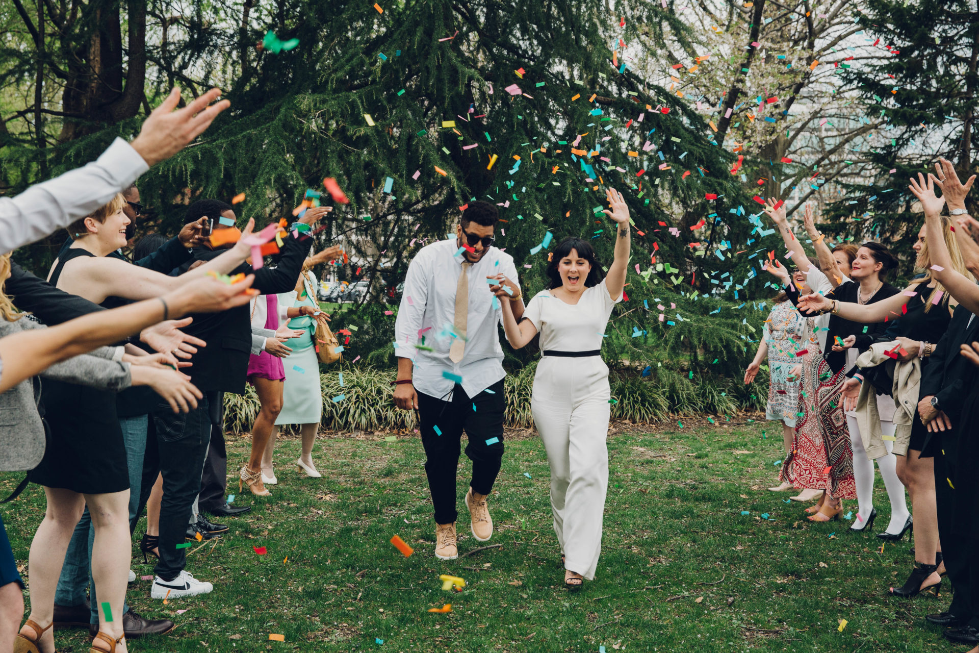 a couple walks in a park underneath a shower of colorful ticker tape confetti, thrown joyfully by friends and relations. Rachel Couch for Pop! Wed Co.