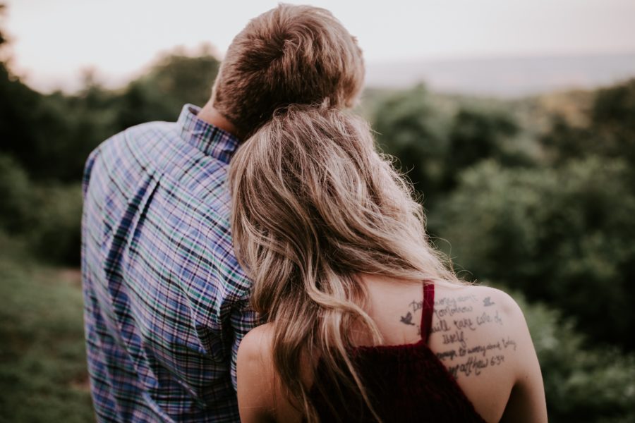 A couple looking out over a lush wooded area, with their backs to you, heads leaned together.