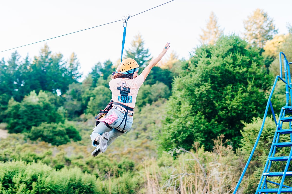 a woman in a pink tee shirt zip lining, with a pink bandana waving from her pocket at the compact summer camp in la Honda, CA