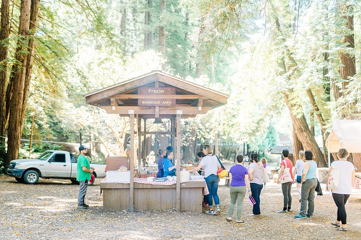 Women lined up to get burgers and hot dogs from a smokey grill stand in a clearing among the redwoods at the compact summer camp in la Honda, CA