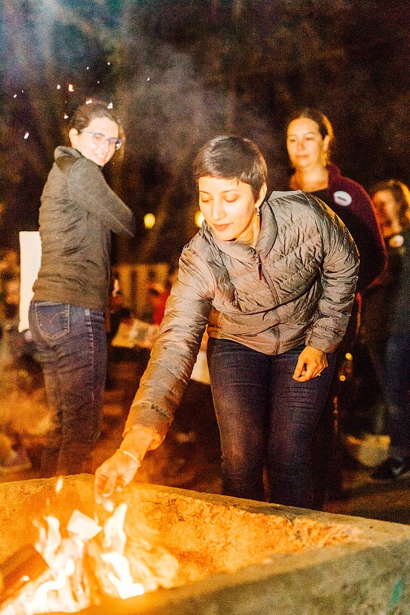 a woman, throwing a piece of paper into the fire as other women look on at the compact summer camp in la Honda, CA