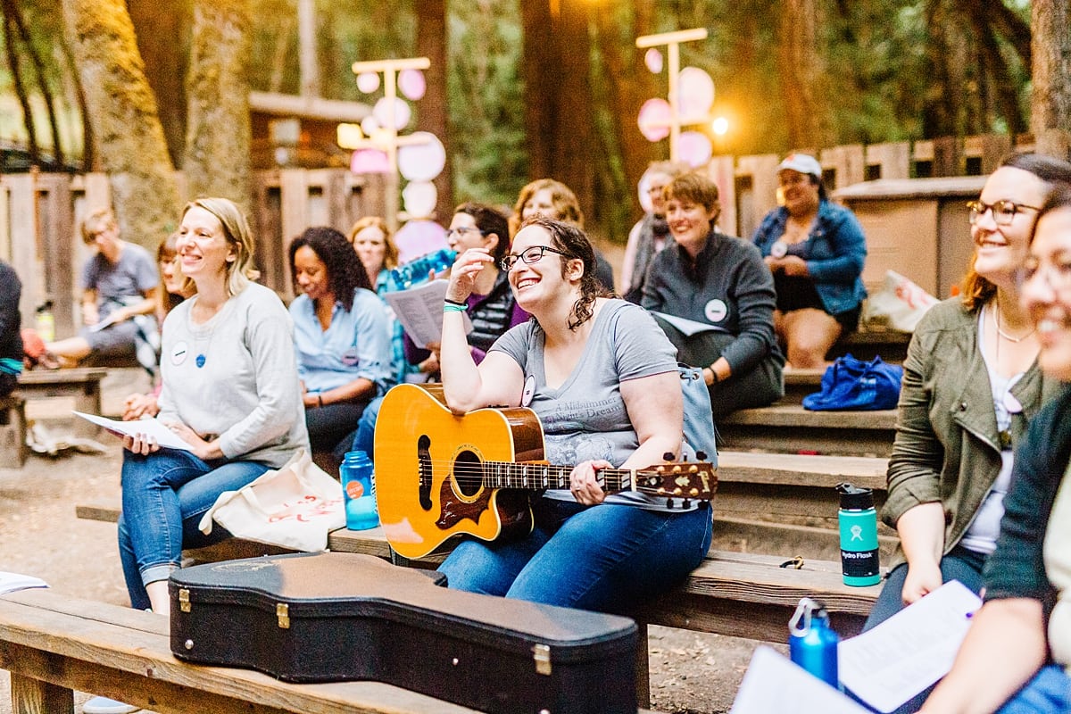 a woman smiling, holding a guitar on a bench among a group of women on benches at the compact summer camp in la Honda, CA