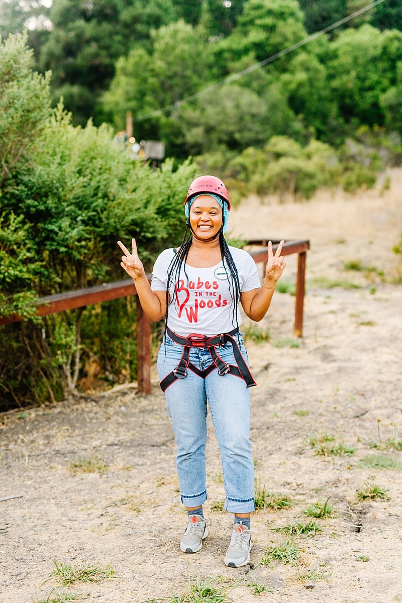 A woman with long braids wearing a babes in the woods tee shirt and zip lining harness and helmet, smiling and throwing peace signs amid the redwoods at the compact summer camp in la Honda, CA