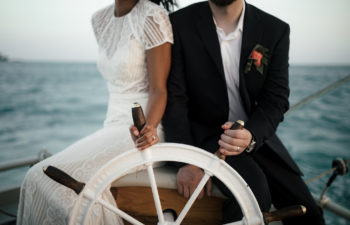 a bride and groom sit on a boat, holding the wheel together, on the sea