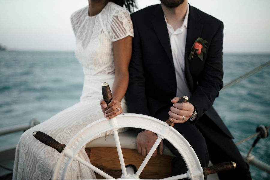 a bride and groom sit on a boat, holding the wheel together, on the sea