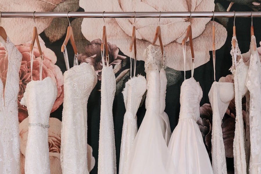 white wedding dresses hang on wooden hangers from a garment rack in front of an oversize floral wall paper at Our Story Bridal