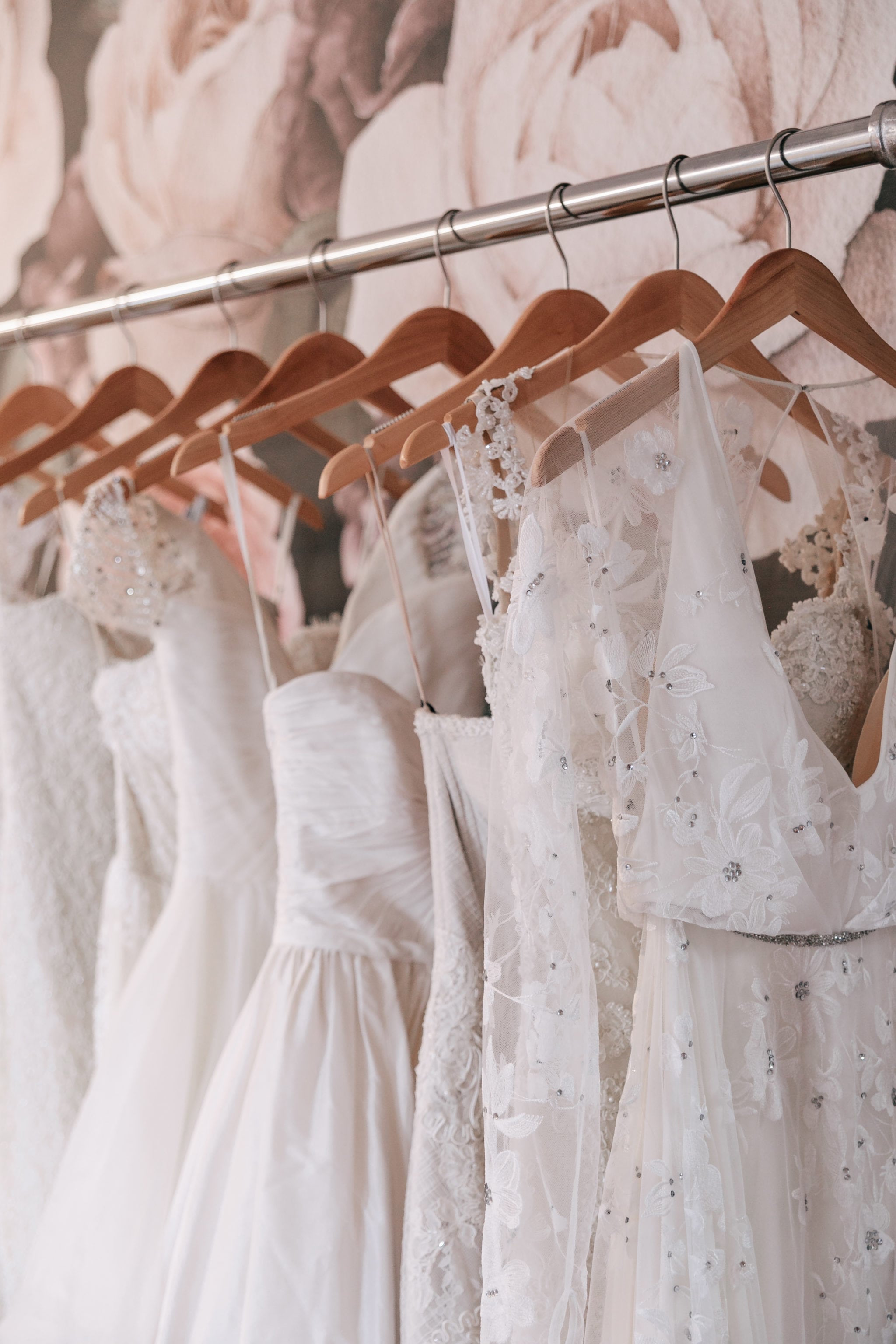close up of white wedding dresses hang on wooden hangers from a garment rack in front of an oversize floral wall paper at Our Story Bridal