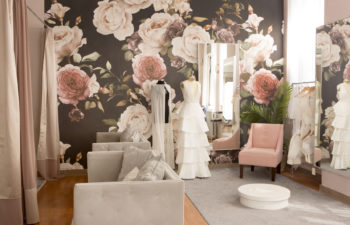 A wedding dress and sheer cape hang on two dress forms in a showroom/fitting room with oversize floral wall paper and modern grey couches with a millennial pink armchair and a mirror at Our Story Bridal