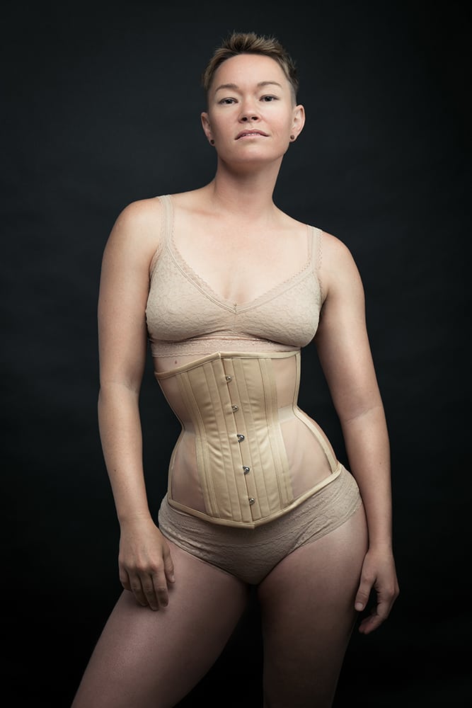 a gender nonconforming person wearing a skin-tone corset from Dark Garden with skin-tone bralette and knickers