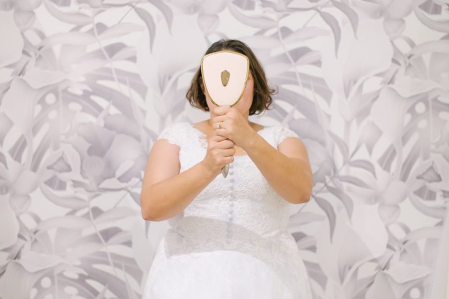 A plus size woman with chin-length dark hair wearing a wedding dress stands in front of a faded tropical leaf print papered wall, holding a hand mirror in front of her face