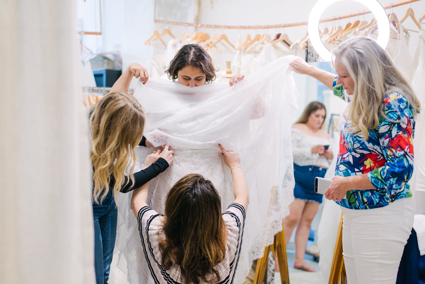 A long hair brunette woman in a striped blouse, a long hair blonde woman and a woman with long grey hair helping plus size woman with short grey hair fix her lace wedding dress in front of plus size woman in short jean skirt and lace blouse on the phone and wedding dresses hanging on wooden hangers 
