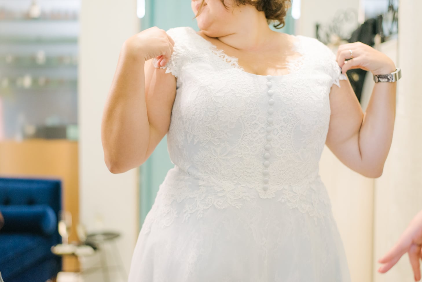 A plus size woman with short brunette hair touches her shoulders while trying on lace wedding dress with buttons down the middle
