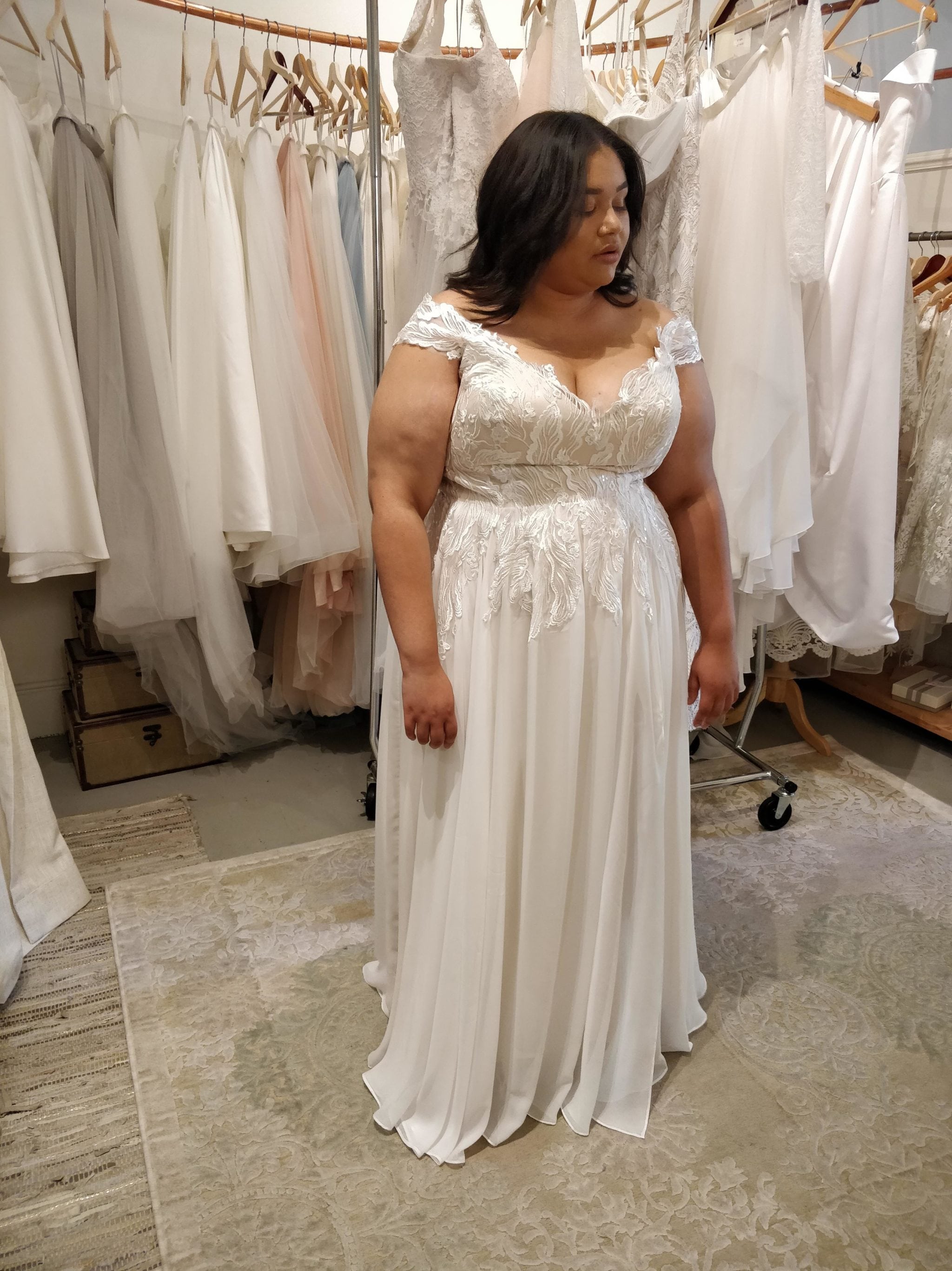 woman with straight dark hair wearing off the shoulder plus size lace wedding dress gown standing in front of other lace wedding dresses hanging from brown wooden hangers