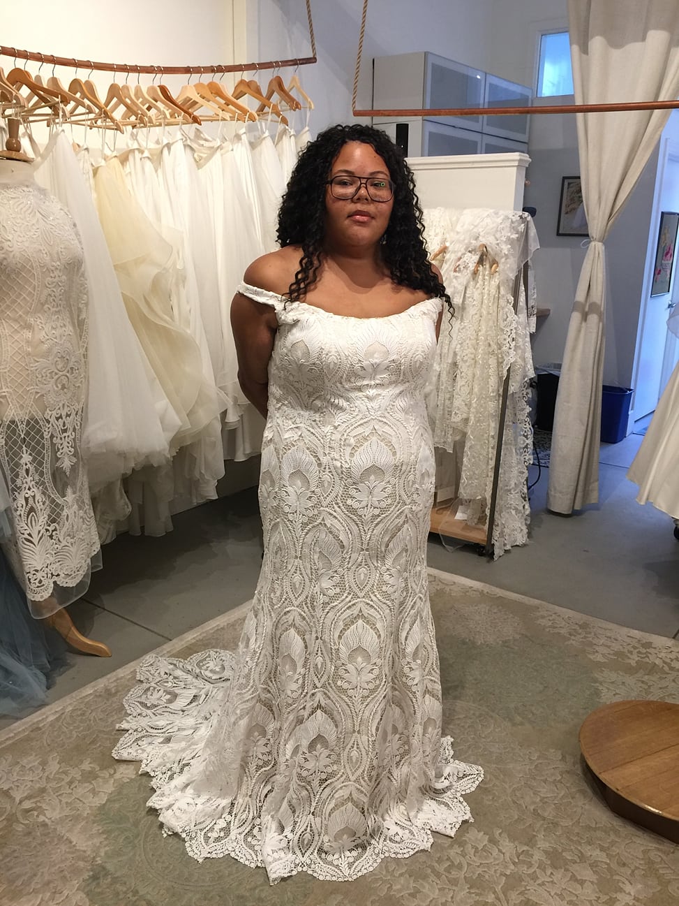 woman wearing glasses with long curly black hair wear long off the shoulder lace wedding dress standing in front of other lace wedding dresses hanging on brown wooden hangers