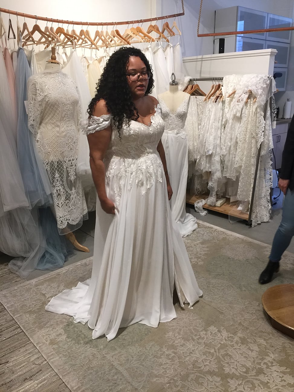 woman wearing glasses with black curly hair wears off the shoulder plus size lace bridal gown standing in front of other lace bridal dresses hanging on wooden hangers 