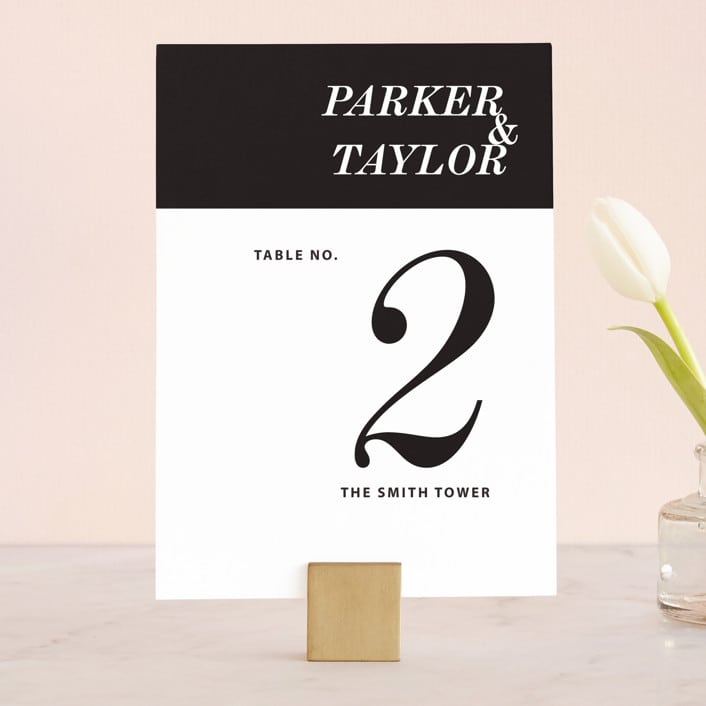 a modern black and white table number from minted styled on a gold place card holder with a single white tulip out of frame
