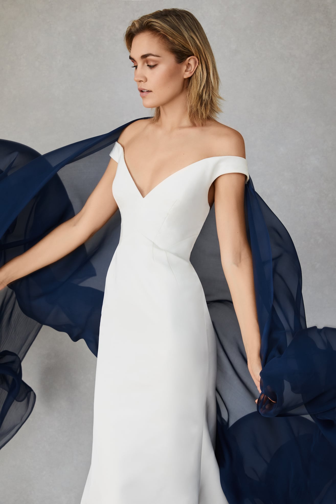 A woman wearing a simple off the shoulder deep v neck wedding dress with a column skirt from Our Story Bridal. She holds a navy sheer wrap that is billowing behind her.