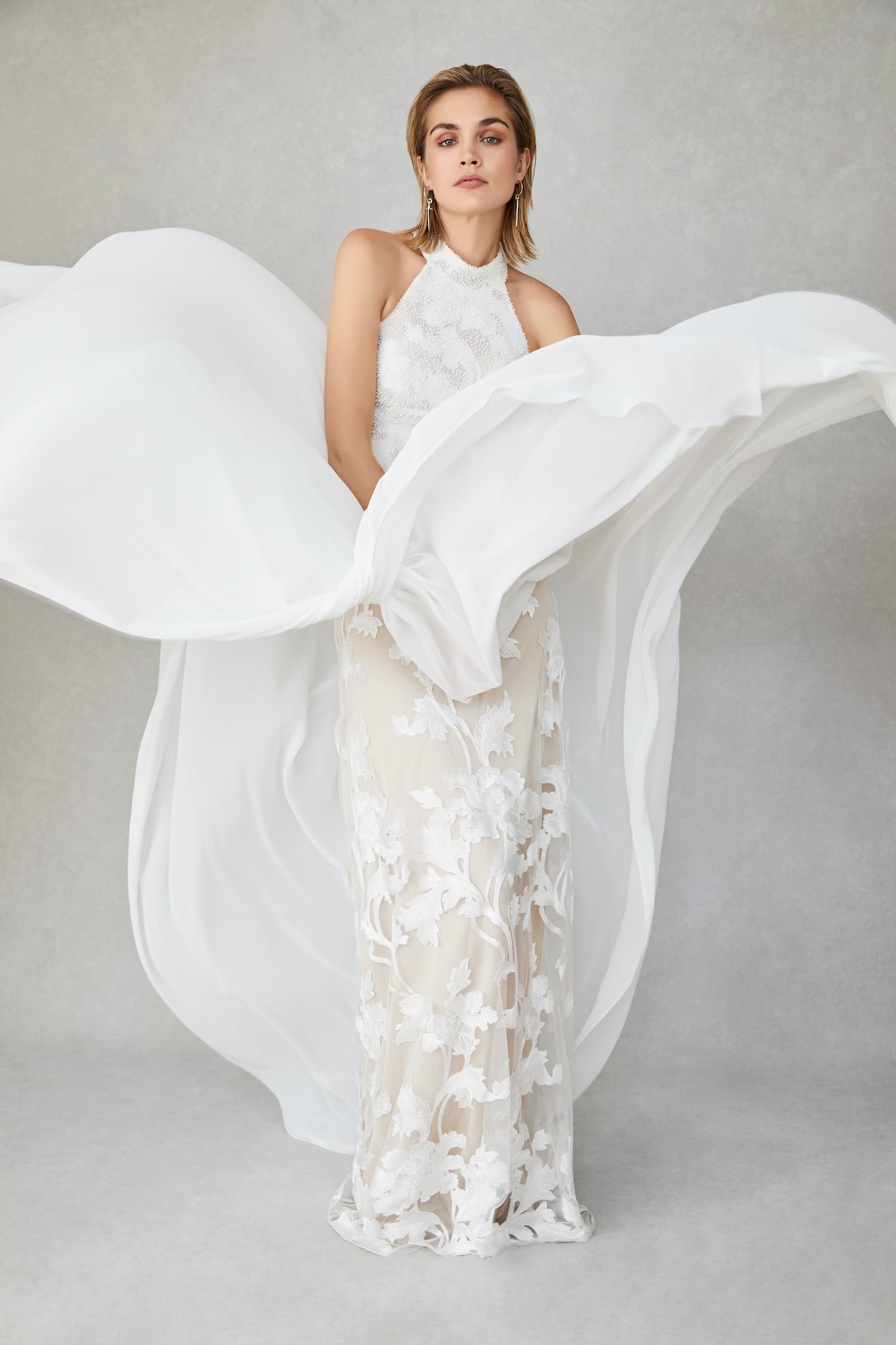 A woman wears a halter neck white wedding dress with a gauzy overlay on top of a sheer dress with botanical style appliqués from Our Story Bridal. Her top overlay skirt billows around her 