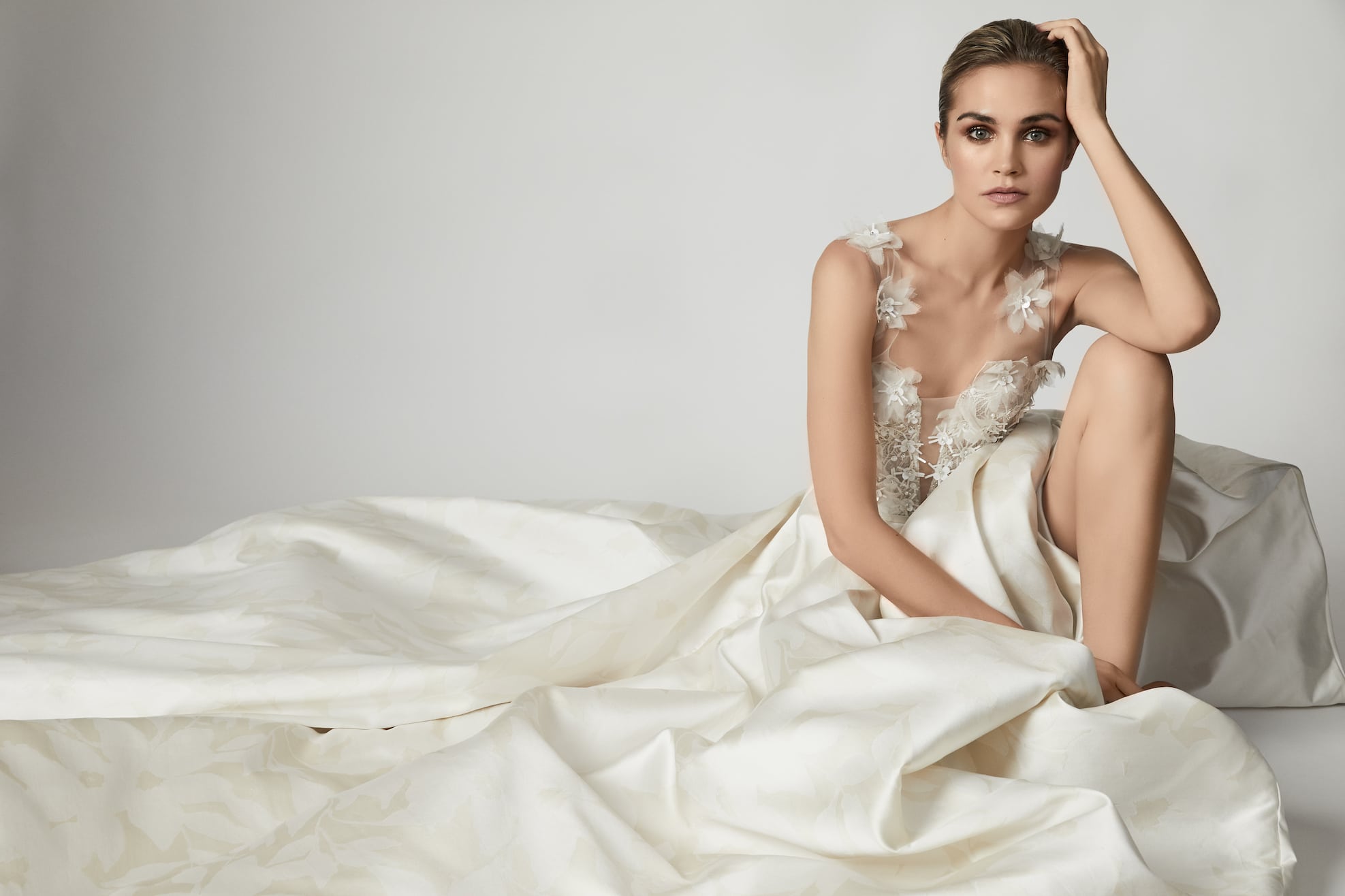 A woman sits on the ground in a flowing off-white gown from Our Story Bridal with floral appliqués on the bodice and illusion straps