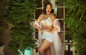 A woman in a vintage pin-up style white lingerie set that may be worn as wedding lingerie and sheer pink robe stands with her hands on her waist in a garden by French doors surrounded by green foliage and near a table with a green watering can