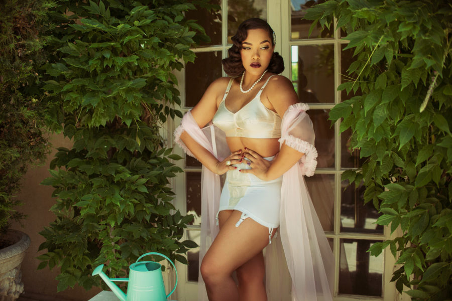 A woman in a vintage pin-up style white lingerie set that may be worn as wedding lingerie and sheer pink robe stands with her hands on her waist in a garden by French doors surrounded by green foliage and near a table with a green watering can