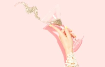 A woman's hand tosses the gold glitter contents of a martini glass in the air in front of a pink wall.