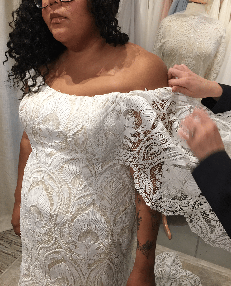 woman wearing glasses with freckles and long dark curly hair wearing long lace wedding dress while strap is being adjusted by a woman wearing black long sleeves 