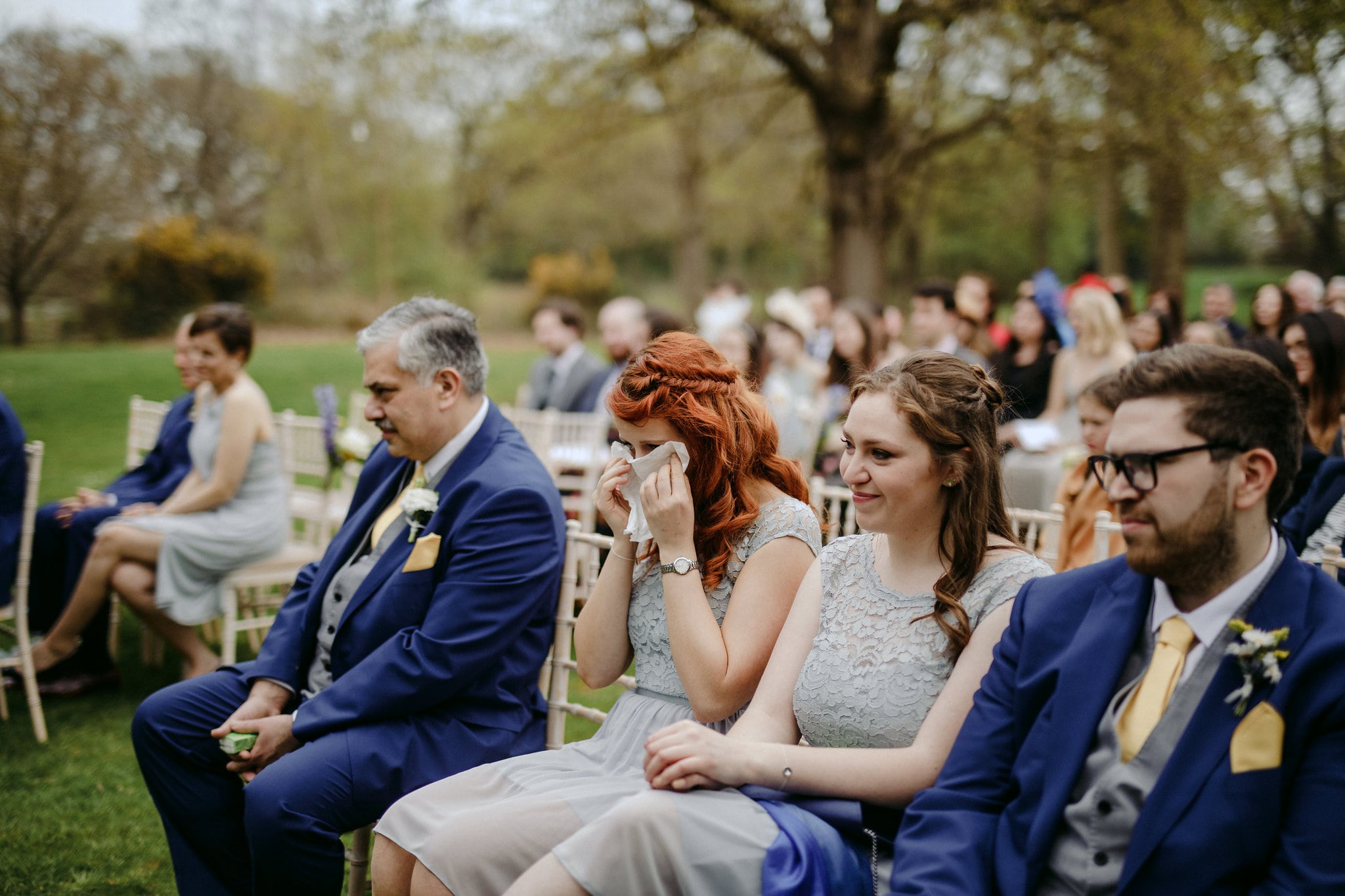 a redheaded bridesmaid wipes tears from both eyes with a handkerchief as she sits among the wedding guests