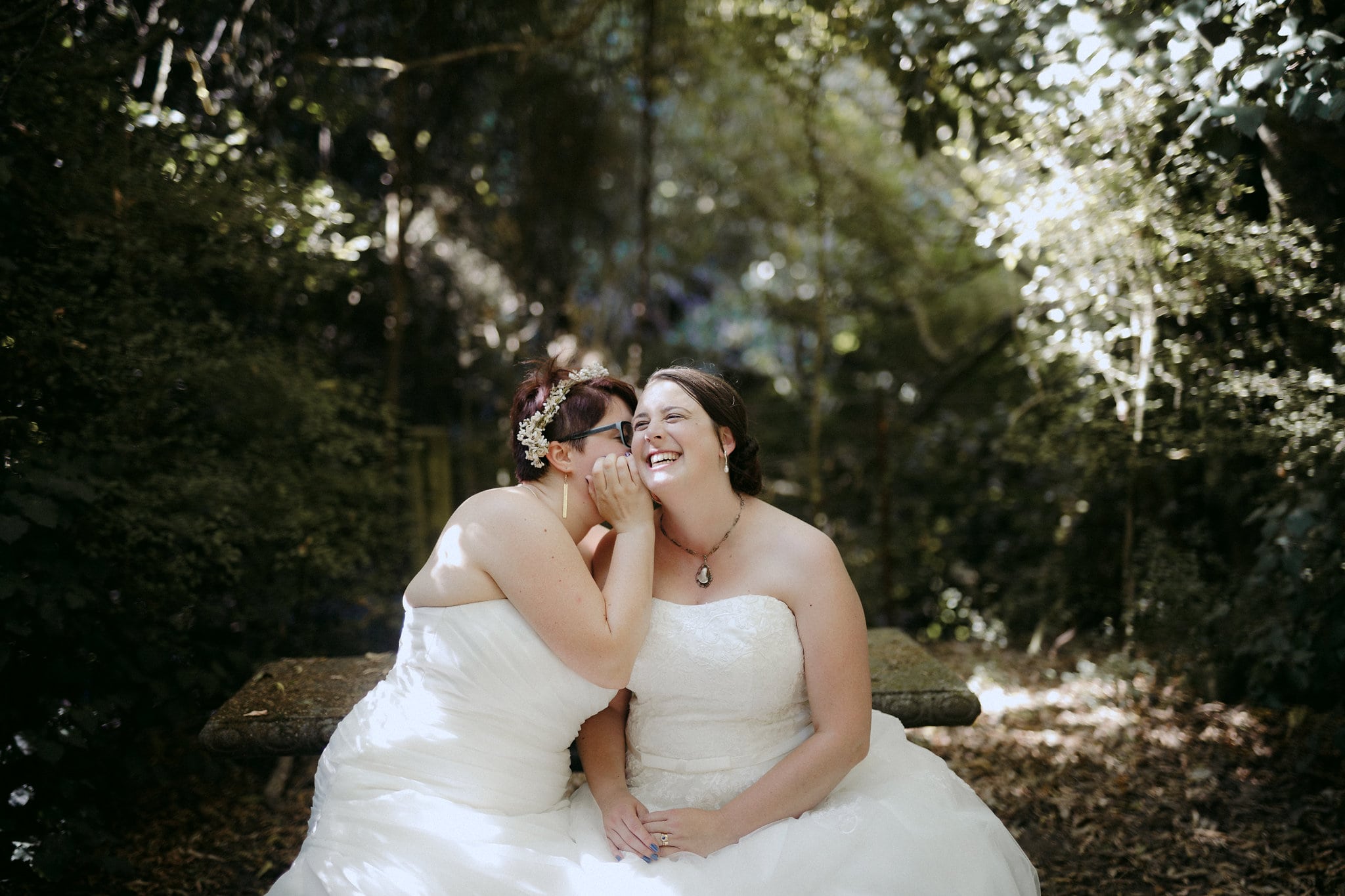 two brides sit on a stone bench among the trees whispering and laughing together