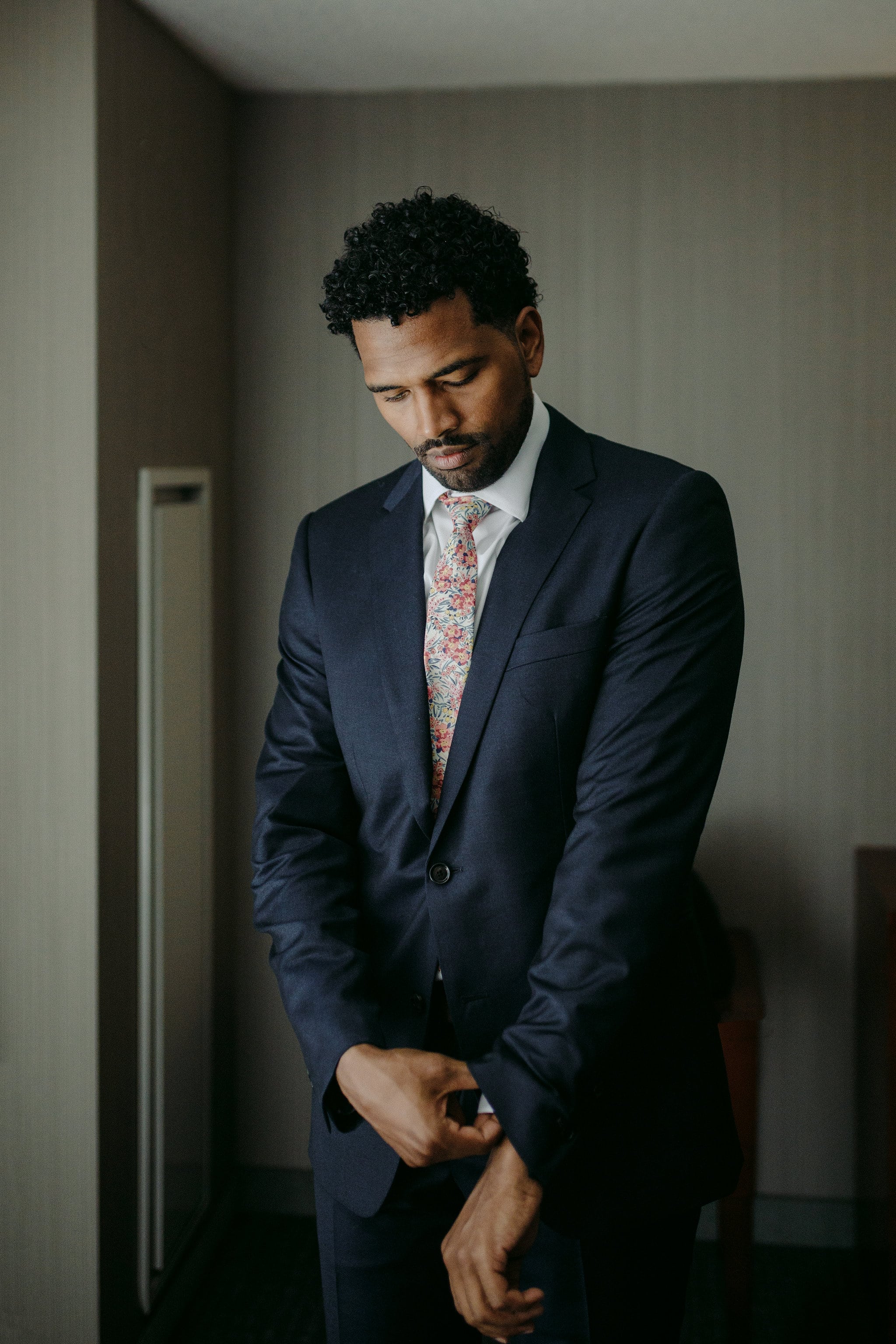 a groom in a dark blue suit with a floral tie adjusts his cuff as he gets ready for his wedding
