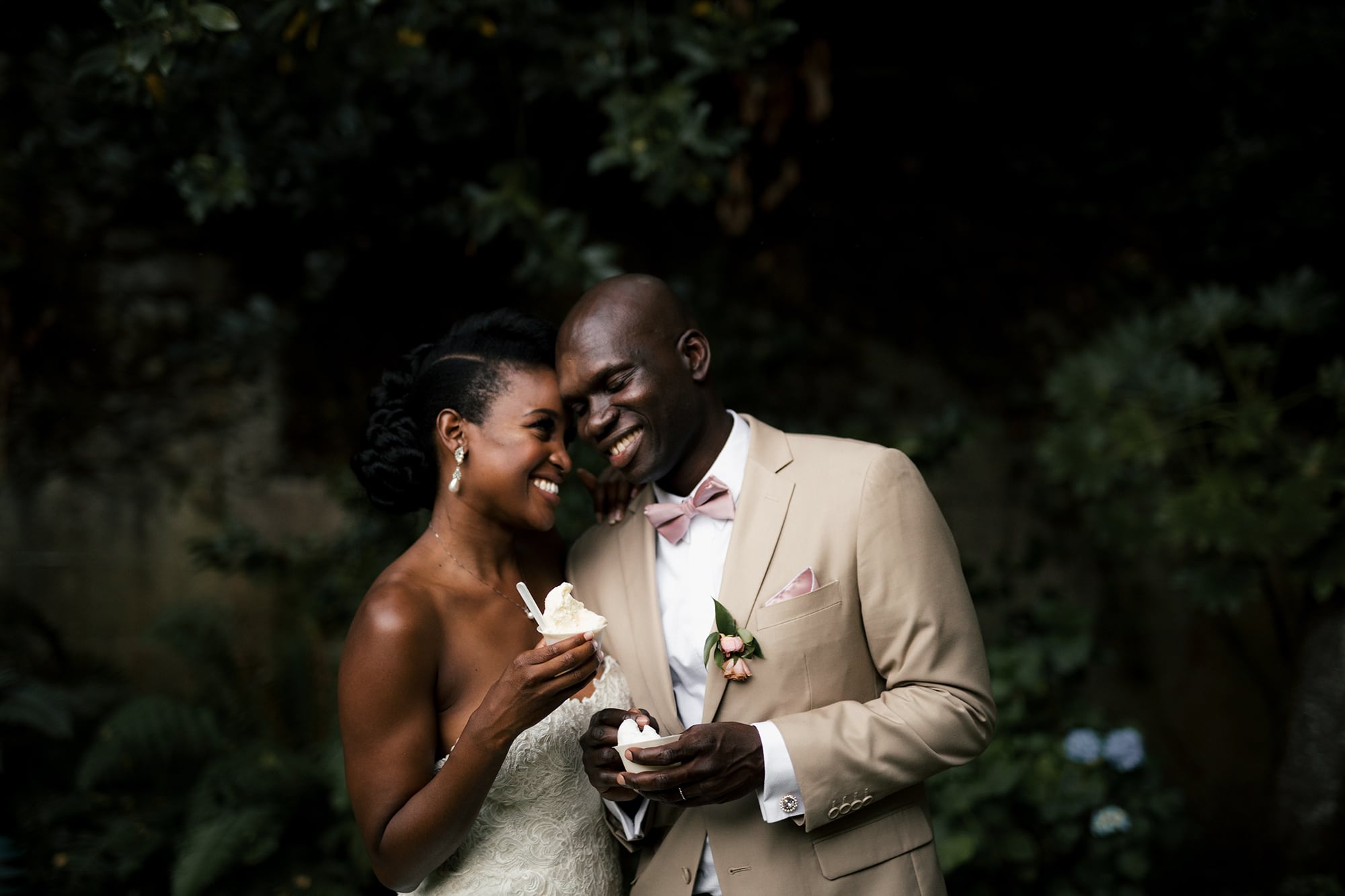 A couple in wedding clothes smiles and leans into one another as they hold cups of vanilla soft serve